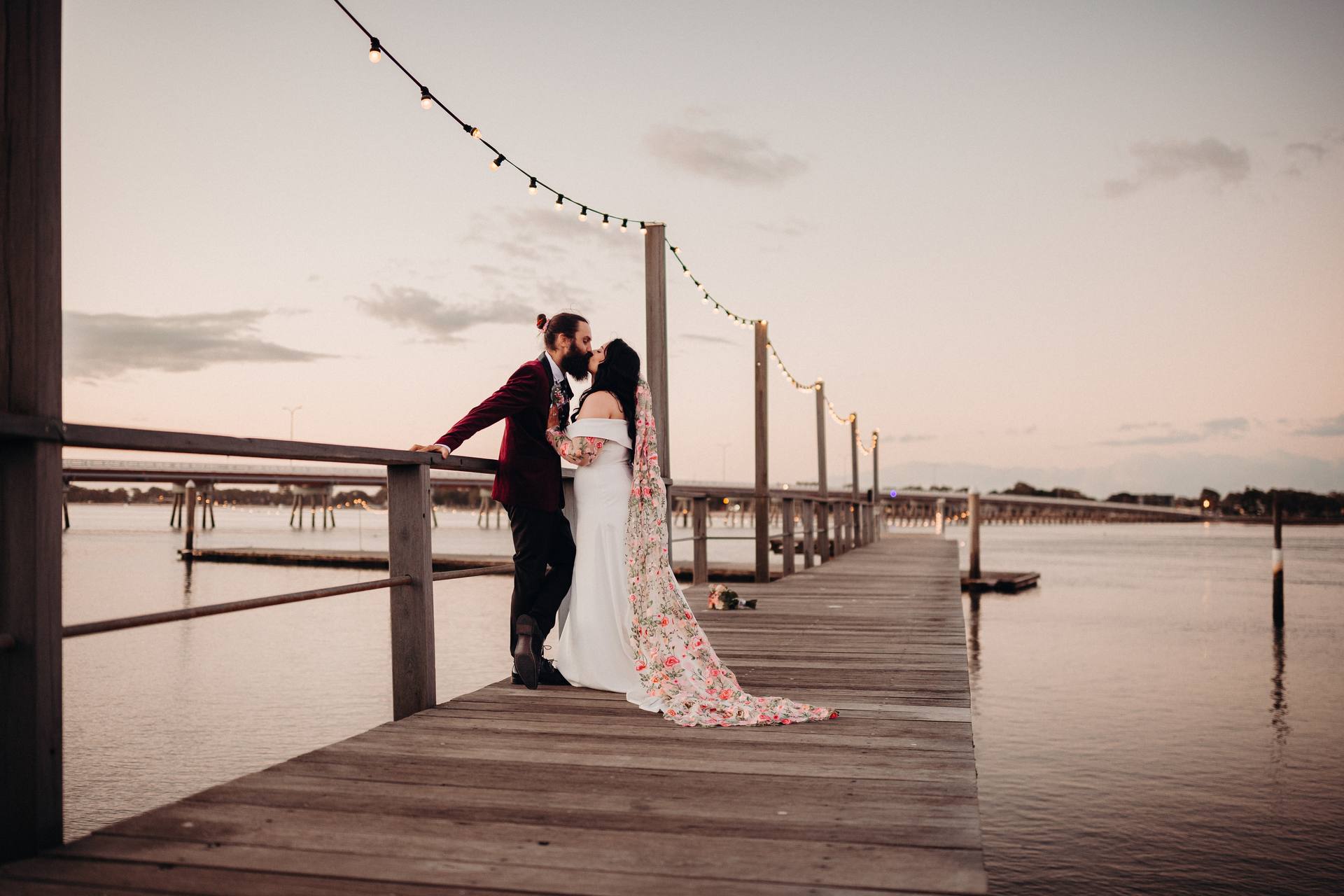 The bride and groom share a kiss on a wooden pier at Sandstone Point Hotel during their couple portraits session at sunset. The bride wears a white gown with a floral-patterned veil, and the groom wears a dark suit jacket with black pants. String lights hang above the pier extending into the water.