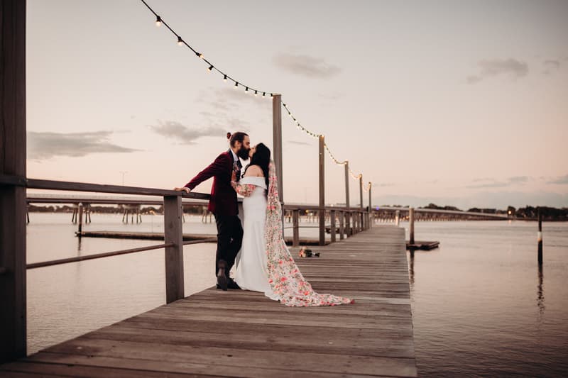 The bride and groom share a kiss on a wooden pier at Sandstone Point Hotel during their couple portraits session at sunset. The bride wears a white gown with a floral-patterned veil, and the groom wears a dark suit jacket with black pants. String lights hang above the pier extending into the water.
