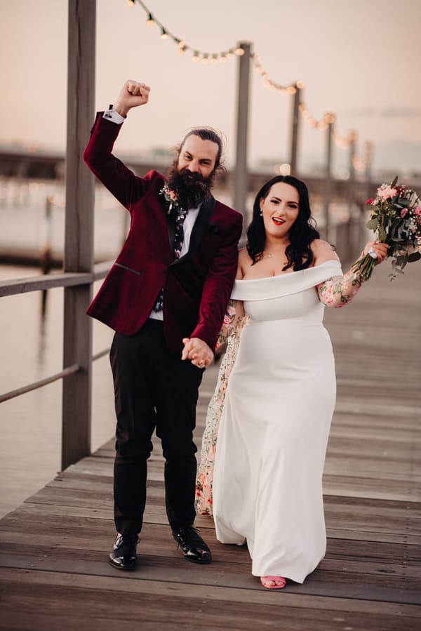 The bride Mindy in a white off-shoulder gown holding a bouquet and the groom David in a burgundy jacket and black pants hold hands and pose on a wooden pier at Sandstone Point Hotel with string lights in the background.