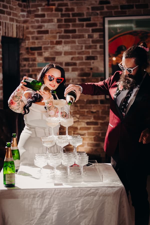 The bride Mindy and the groom David, both wearing heart-shaped sunglasses, pour champagne into a pyramid of coupe glasses at the Sandstone Point Hotel — Cellar during the reception stage.