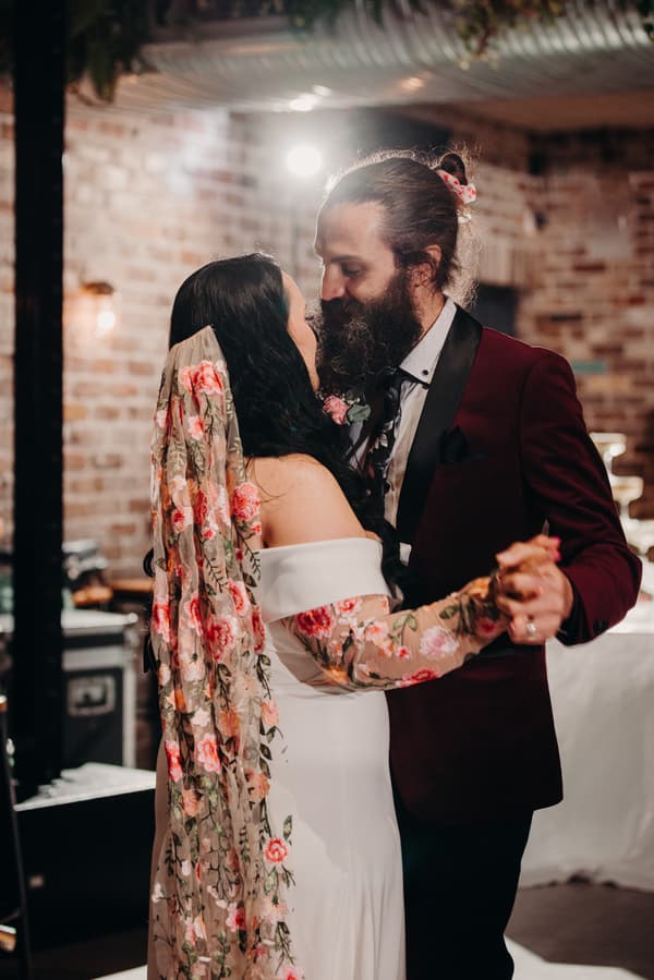 The bride Mindy and groom David share a close dance on the reception stage at Sandstone Point Hotel — Cellar. Mindy wears an off-shoulder white dress with a floral veil, and David wears a burgundy suit jacket with a black lapel and a floral boutonniere.