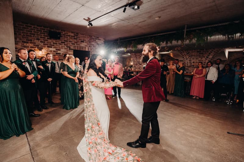 Bride Mindy and groom David dance together on the reception stage at Sandstone Point Hotel — Cellar, surrounded by guests and bridal party members watching.