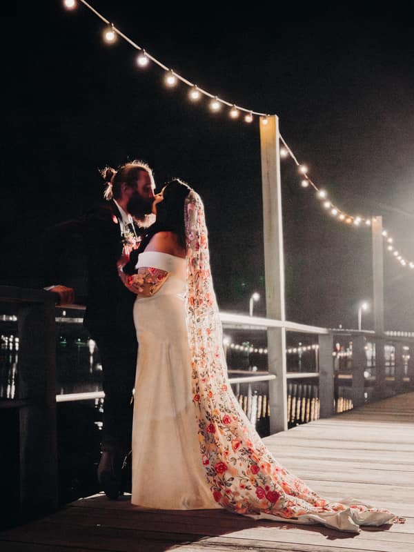 The bride and groom share a kiss on a wooden pier decorated with string lights at night at Sandstone Point Hotel — Cellar.