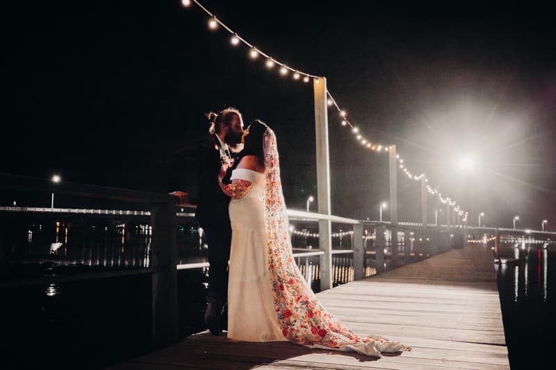 The bride and groom share a kiss on a wooden pier decorated with string lights at night at Sandstone Point Hotel — Cellar.