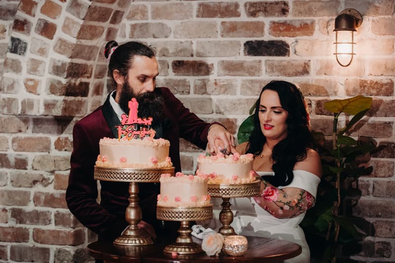 Bride Mindy and groom David cut the wedding cake on the reception stage at Sandstone Point Hotel — Cellar, with three cakes displayed on ornate cake stands against a brick wall backdrop.