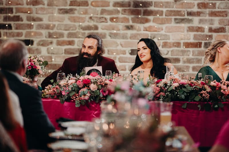 The bride Mindy and groom David sit at a reception table decorated with floral arrangements and a pink tablecloth at Sandstone Point Hotel — Cellar, facing guests during the reception.