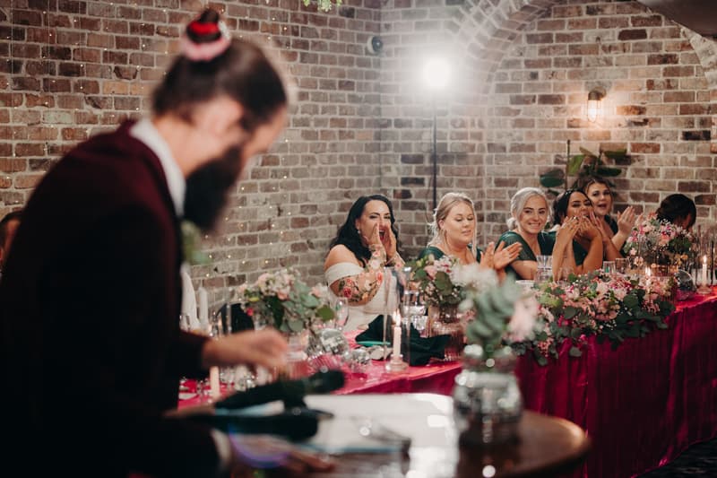 The bride and bridesmaids sit at a long table decorated with flowers and candles at Sandstone Point Hotel — Cellar, applauding a bearded man in a burgundy jacket who is speaking or performing in the foreground.