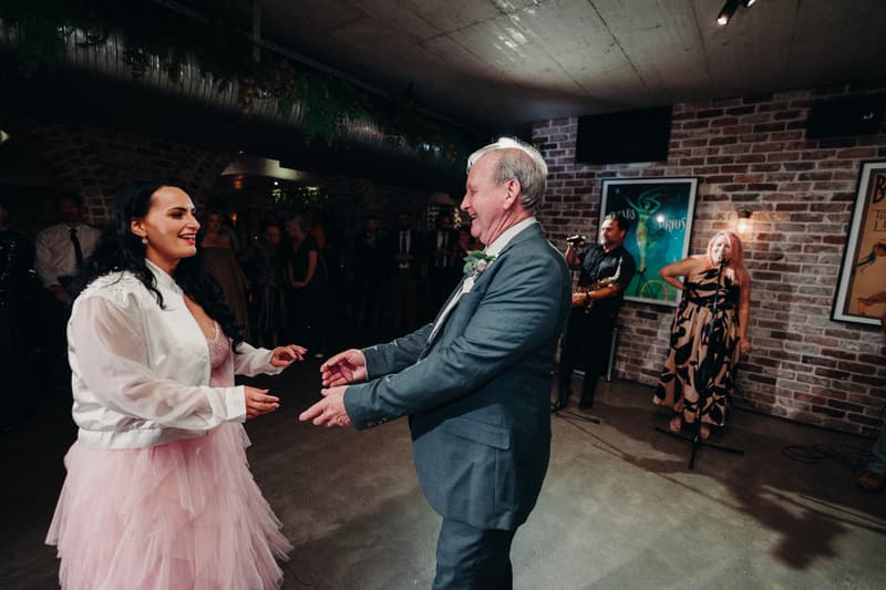 The bride dances with an older man, likely her father, at the Sandstone Point Hotel — Cellar during the wedding reception. In the background, a woman sings into a microphone while a man plays a saxophone, with guests watching.