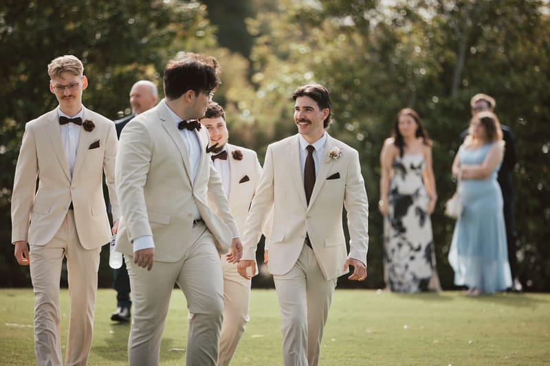 Joel and groomsmen walk outdoors on grass, dressed in cream suits with brown bow ties and boutonnieres, with guests visible in the background. Sunset wedding portraits Brisbane by She Said Yes.