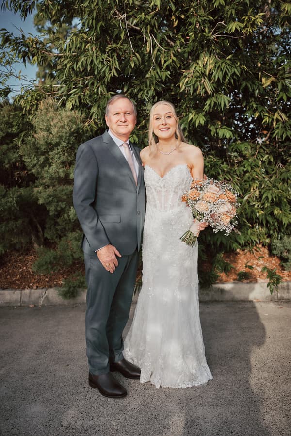 The bride Morgan stands next to an older man, likely her father, outdoors at Sandstone Point Hotel — The Pavilion, holding a bouquet of flowers.