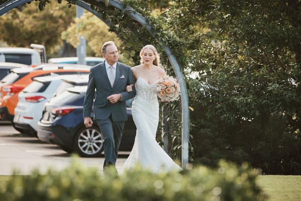 The bride Morgan walks arm-in-arm with an older man, likely her father, under a metal archway near a parking lot. Photo and video services by She Said Yes.