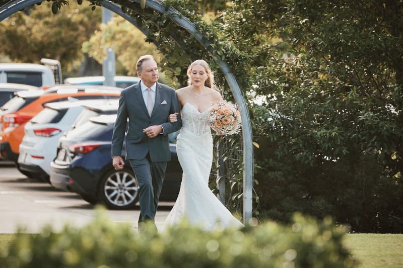 The bride Morgan walks arm-in-arm with an older man, likely her father, under a metal archway near a parking lot at Sandstone Point Hotel — The Pavilion.