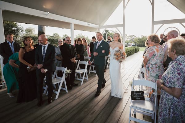 The bride Morgan walks down the aisle arm-in-arm with a man, likely her father,, while guests stand on either side watching. Brisbane videographer by She Said Yes.