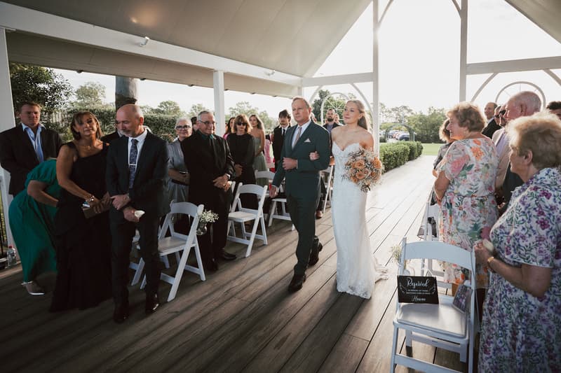 The bride Morgan walks down the aisle arm-in-arm with a man, likely her father, at Sandstone Point Hotel — The Pavilion, while guests stand on either side watching.