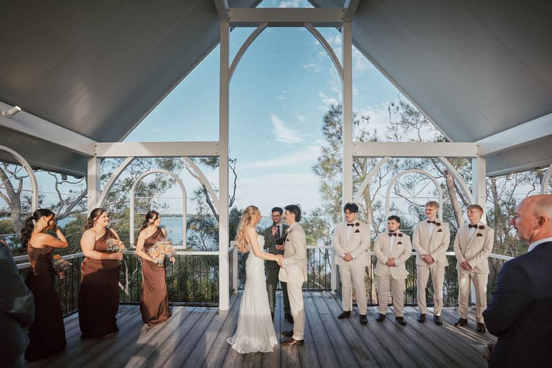 Bride Morgan and groom Joel hold hands facing each other during their wedding ceremony at Sandstone Point Hotel — The Pavilion, with bridesmaids in brown dresses and groomsmen in cream suits standing on either side and an officiant behind them.