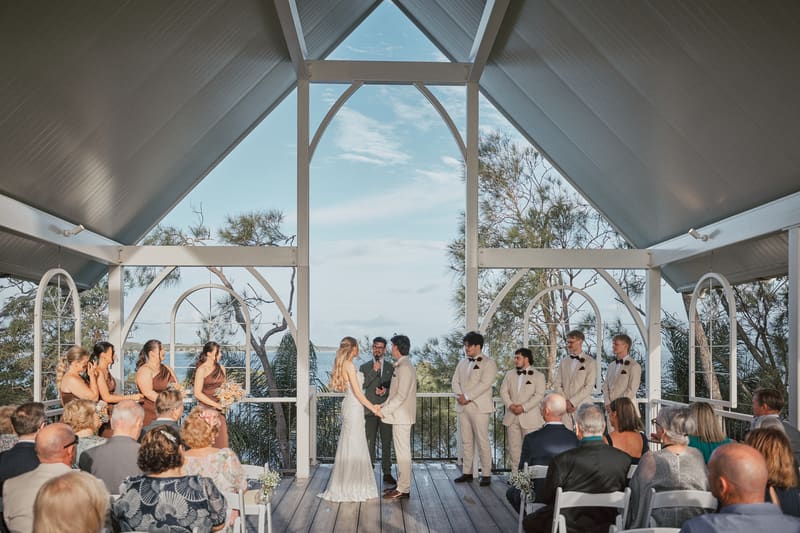 Bride Morgan and groom Joel stand holding hands at the altar with the officiant, surrounded by bridesmaids in brown dresses on the left and groomsmen in beige suits on the right, with seated guests watching the ceremony. School holiday wedding planning by She Said Yes.