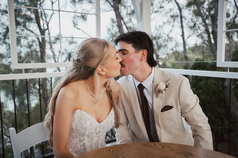 The bride Morgan and groom Joel share a kiss while seated at a wooden table at Sandstone Point Hotel — The Pavilion, with trees visible outside the window behind them.