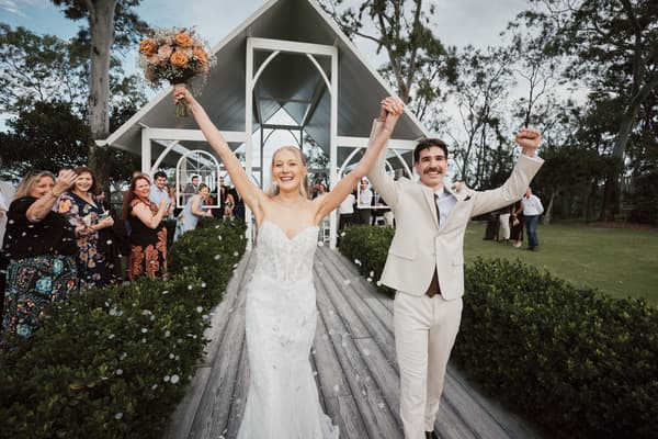 Bride Morgan and groom Joel walk down the aisle holding hands with raised arms, guests stand on either side applauding. Brisbane videographer by She Said Yes.