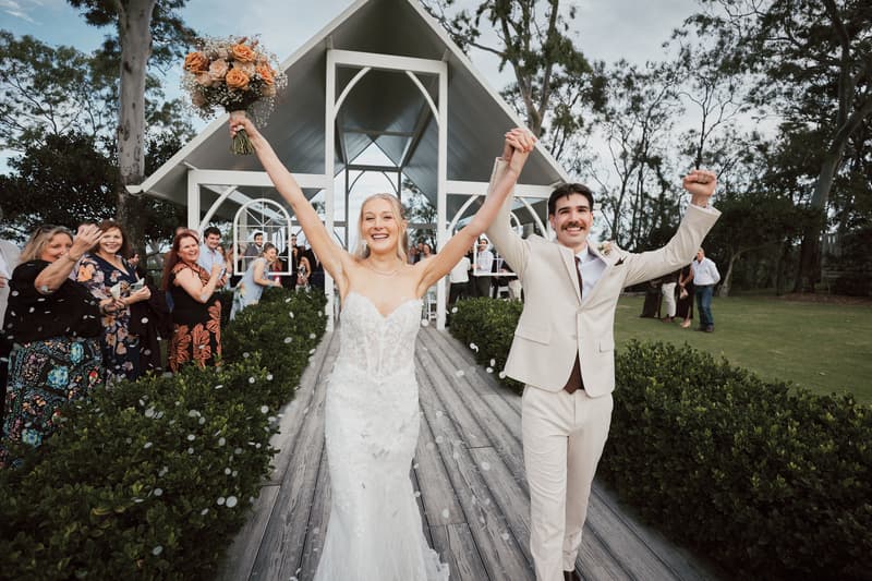 Bride Morgan and groom Joel walk down the aisle holding hands with raised arms at Sandstone Point Hotel — The Pavilion, guests stand on either side applauding.