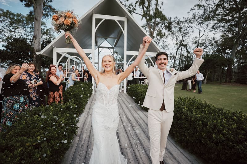 Bride Morgan and groom Joel walk down the aisle holding hands with raised arms, guests stand on either side applauding. Lead follow-up communication issues by She Said Yes.