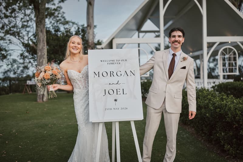 Bride Morgan in a white wedding gown holding a bouquet and groom Joel in a beige suit stand outdoors at Sandstone Point Hotel, posing beside a wedding welcome sign with their names and wedding date.