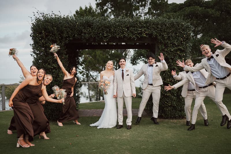 Bride Morgan and groom Joel stand holding hands under a leafy arch at Sandstone Point Hotel, surrounded by bridesmaids in brown dresses and groomsmen in beige suits posing playfully.