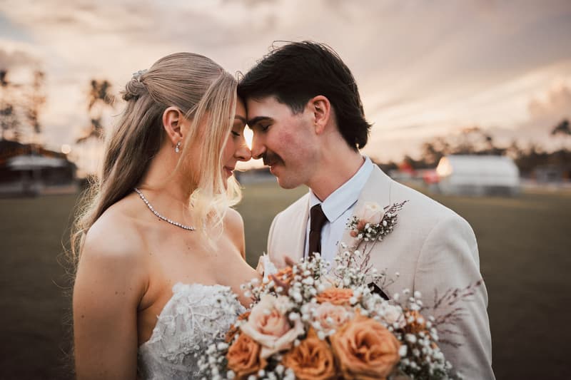 Bride Morgan and groom Joel pose closely together outdoors at Sandstone Point Hotel during their couple portraits session, with Morgan holding a bouquet of peach and white flowers.