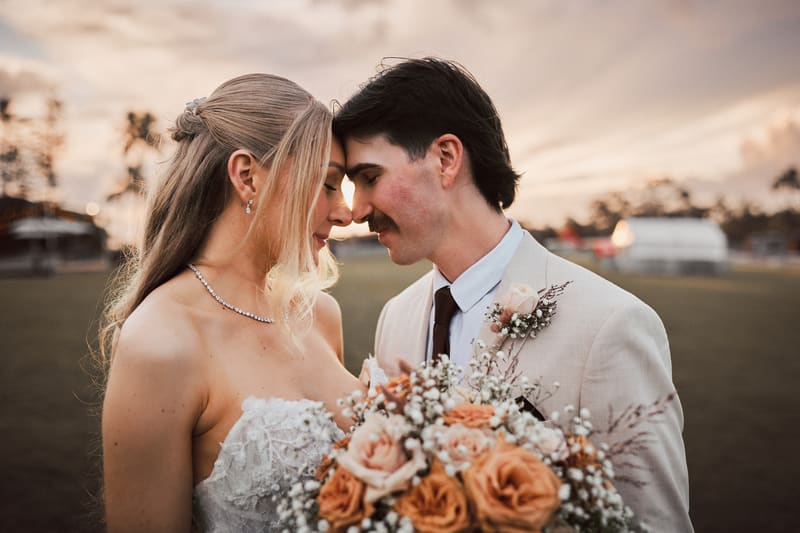 Bride Morgan and groom Joel pose closely together outdoors, with Morgan holding a bouquet of peach and white flowers. Cinematic videographer by She Said Yes.