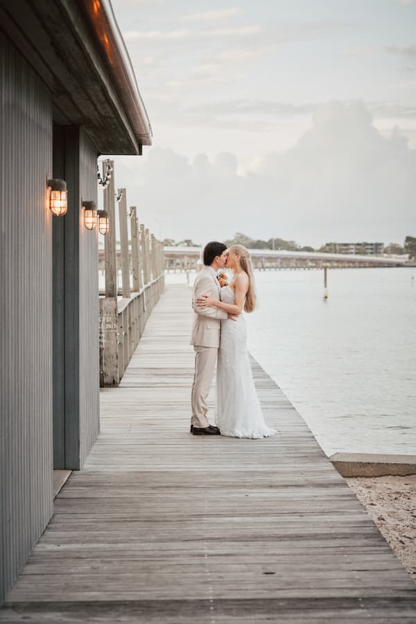 Morgan the bride and Joel the groom embrace and kiss on a wooden pier at Sandstone Point Hotel with water and distant buildings in the background.