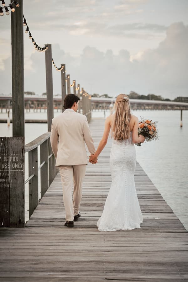 Morgan the bride and Joel the groom walk hand in hand along a wooden pier at Sandstone Point Hotel, with Morgan holding a bouquet of flowers.