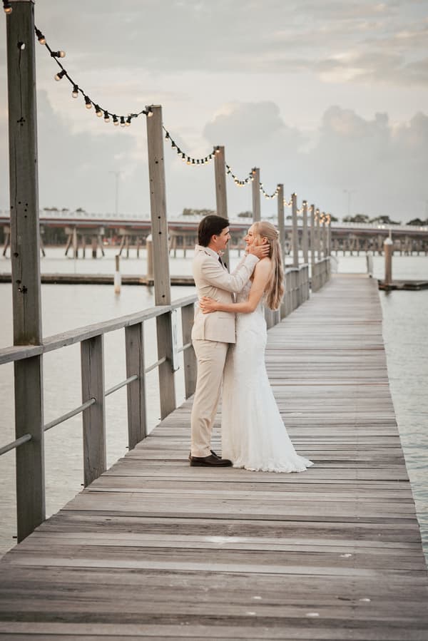 Morgan the bride and Joel the groom embrace on a wooden pier decorated with string lights at Sandstone Point Hotel, with water and a distant bridge visible in the background.