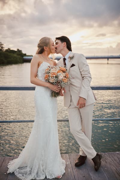 Bride Morgan and groom Joel kiss while standing on a wooden deck by the water. Morgan holds a bouquet of peach and white flowers, and Joel is dressed in a light beige suit. Valentine's Day wedding photography by She Said Yes.