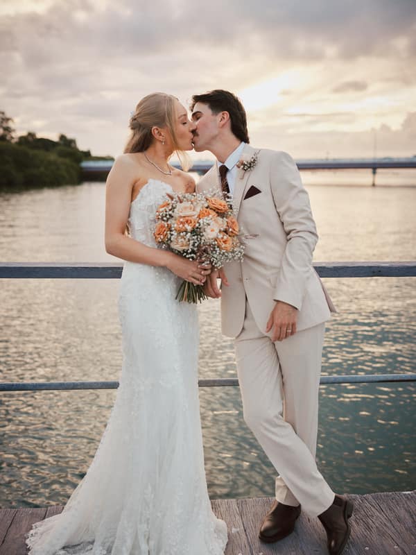 Bride Morgan and groom Joel kiss while standing on a wooden deck by the water at Sandstone Point Hotel during their couple portraits session. Morgan holds a bouquet of peach and white flowers, and Joel is dressed in a light beige suit.