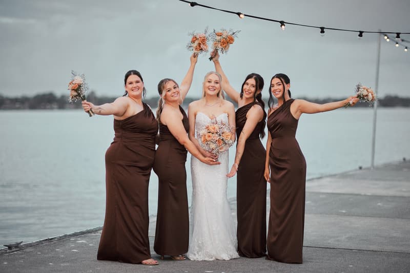 Morgan the bride poses with four bridesmaids on a pier at Sandstone Point Hotel, each holding bouquets with the bride in a white gown and the bridesmaids in matching dark brown dresses.