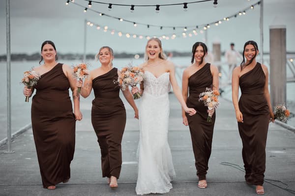 The bride Morgan walks hand in hand with four bridesmaids along a pier, each holding bouquets and smiling under string lights. Elopement photography Brisbane by She Said Yes.