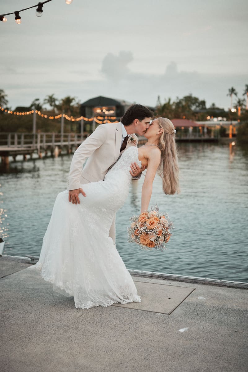 Morgan and Joel share a kiss during their couple portraits, with Joel dipping Morgan who holds a bouquet of peach and white flowers by the waterfront. Engagement celebration by She Said Yes.