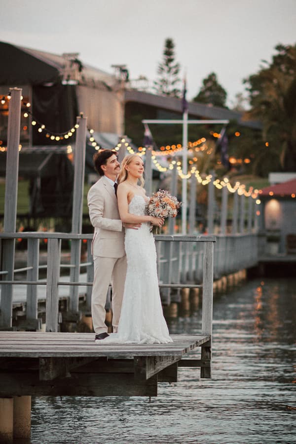 Morgan the bride in a white wedding gown holding a bouquet stands on a wooden pier at Sandstone Point Hotel, embraced from behind by Joel the groom in a light beige suit, with string lights and water visible in the background.
