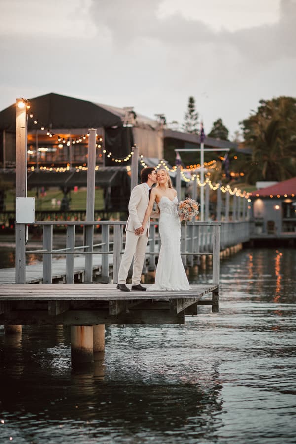Morgan the bride and Joel the groom stand on a wooden pier at Sandstone Point Hotel, with Joel kissing Morgan on the cheek as she holds a bouquet of flowers. String lights and buildings are visible in the background over the water.