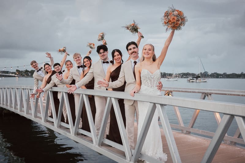 Morgan the bride and Joel the groom pose with their bridal party on a metal bridge over water, with bridesmaids in dark dresses and groomsmen in light suits holding bouquets raised in the air. Wedding photographers Brisbane by She Said Yes.