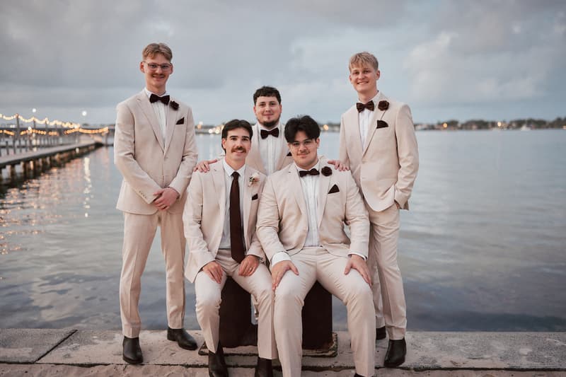 Five groomsmen in beige suits with dark bow ties and boutonnieres pose together by the water at Sandstone Point Hotel during the couple portraits stage.