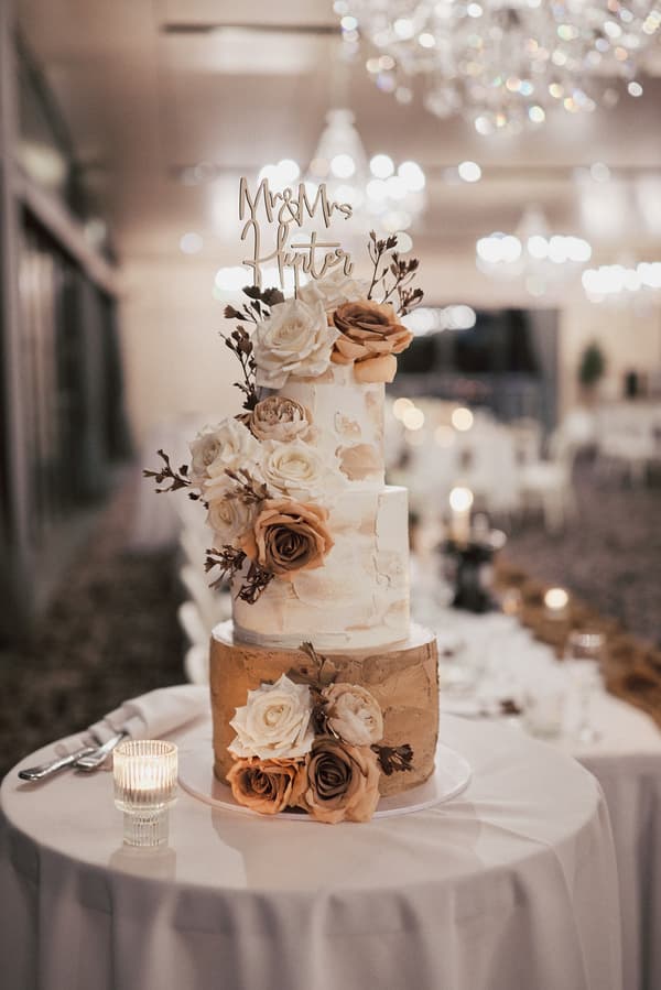 Three-tier wedding cake decorated with white and beige roses and a topper reading 'Mr & Mrs Hunter' on a round table at Sandstone Point Hotel — The Pumicestone Room.