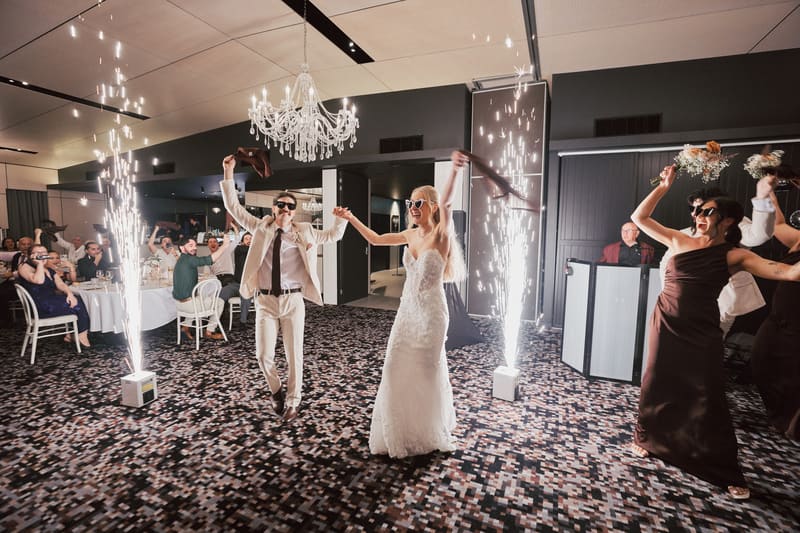 Bride Morgan and groom Joel enter the reception stage, holding hands and wearing sunglasses, with guests seated at tables cheering and bridesmaids holding bouquets and scarves nearby, while indoor sparklers light the scene. Good wedding vows by She Said Yes.