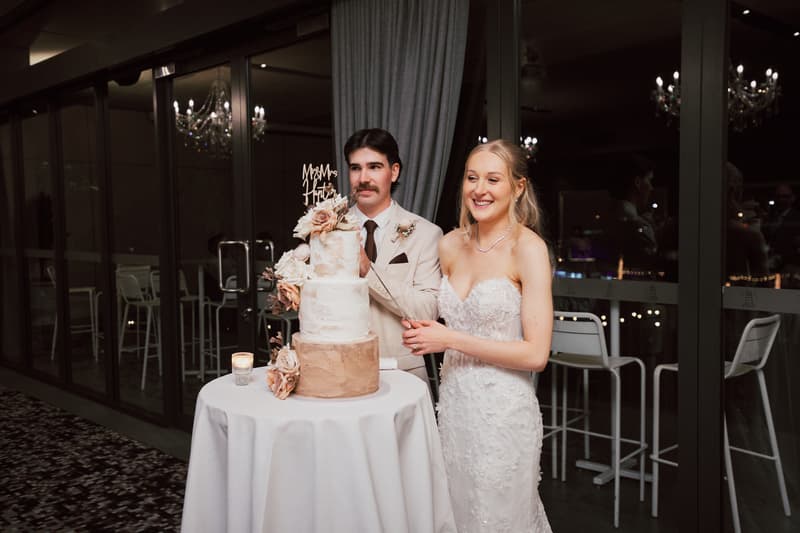 The bride Morgan and groom Joel cut their wedding cake on the reception stage at Sandstone Point Hotel — The Pumicestone Room. The three-tier cake is decorated with flowers and a 'Mr & Mrs' topper.