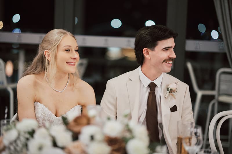 The bride Morgan and groom Joel sit side by side at a table during the reception at Sandstone Point Hotel — The Pumicestone Room, smiling and looking to their right. Morgan wears a strapless white wedding dress and a necklace, while Joel wears a light-colored suit with a dark tie and boutonniere.