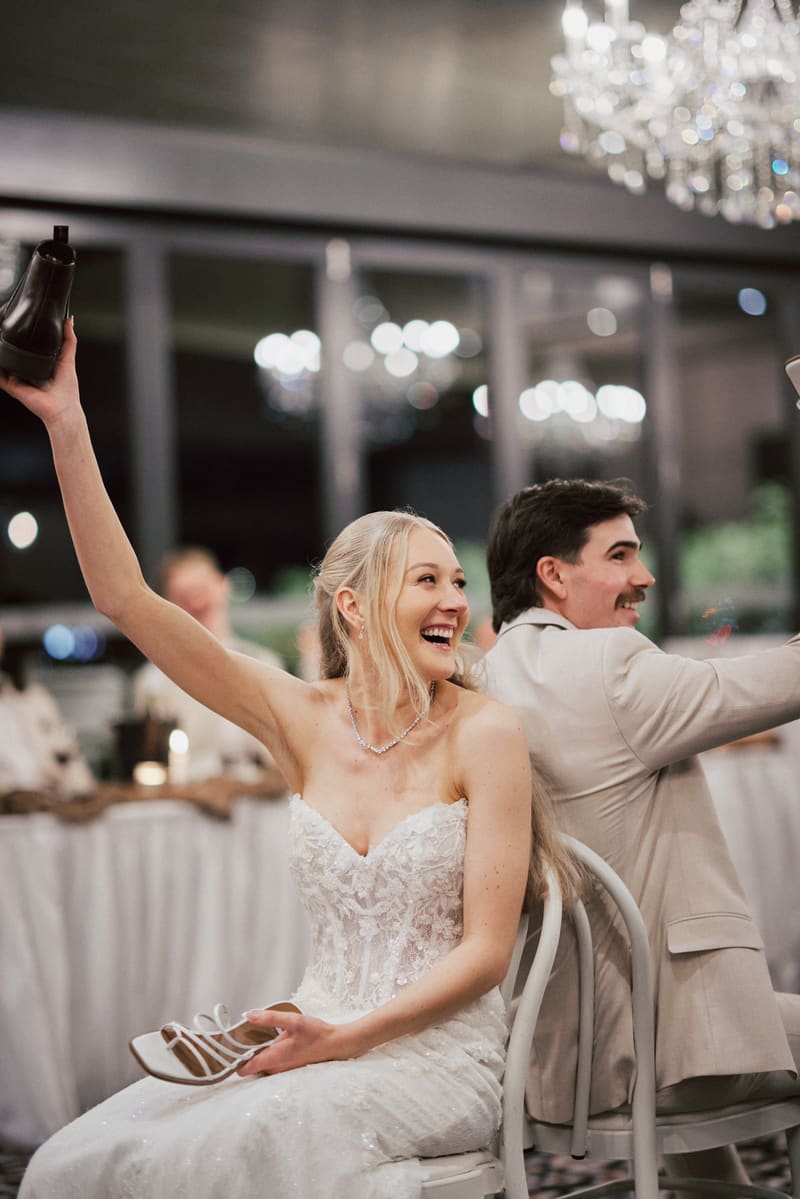 The bride Morgan and groom Joel sit back to back on chairs at the reception in The Pumicestone Room,, each holding up a shoe in a playful wedding shoe game moment. Wedding date changed COVID by She Said Yes.