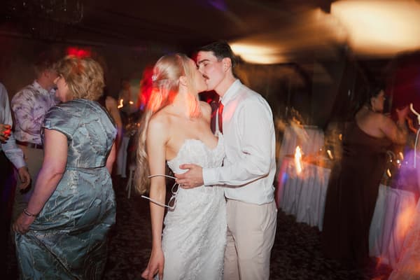 Bride Morgan and groom Joel kiss on the dance floor at the Sandstone Point Hotel — The Pumicestone Room reception, surrounded by guests dancing and mingling.