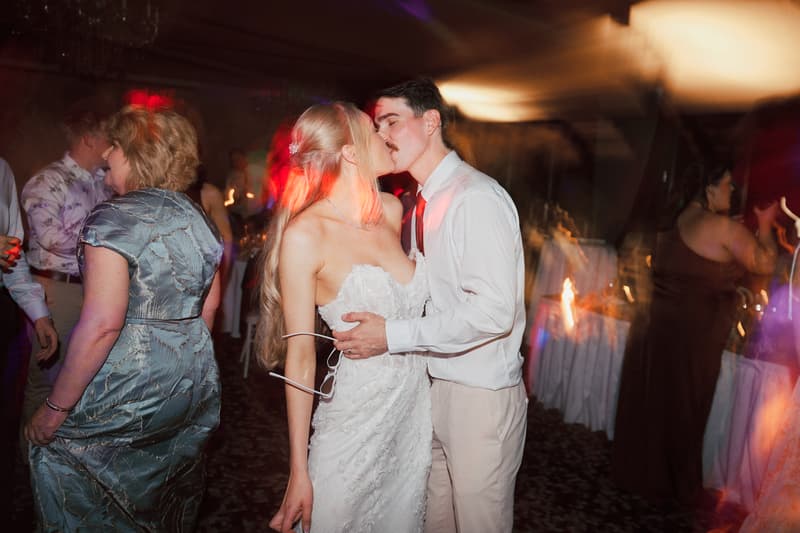 Bride Morgan and groom Joel kiss on the dance floor at the Sandstone Point Hotel — The Pumicestone Room reception, surrounded by guests dancing and mingling.