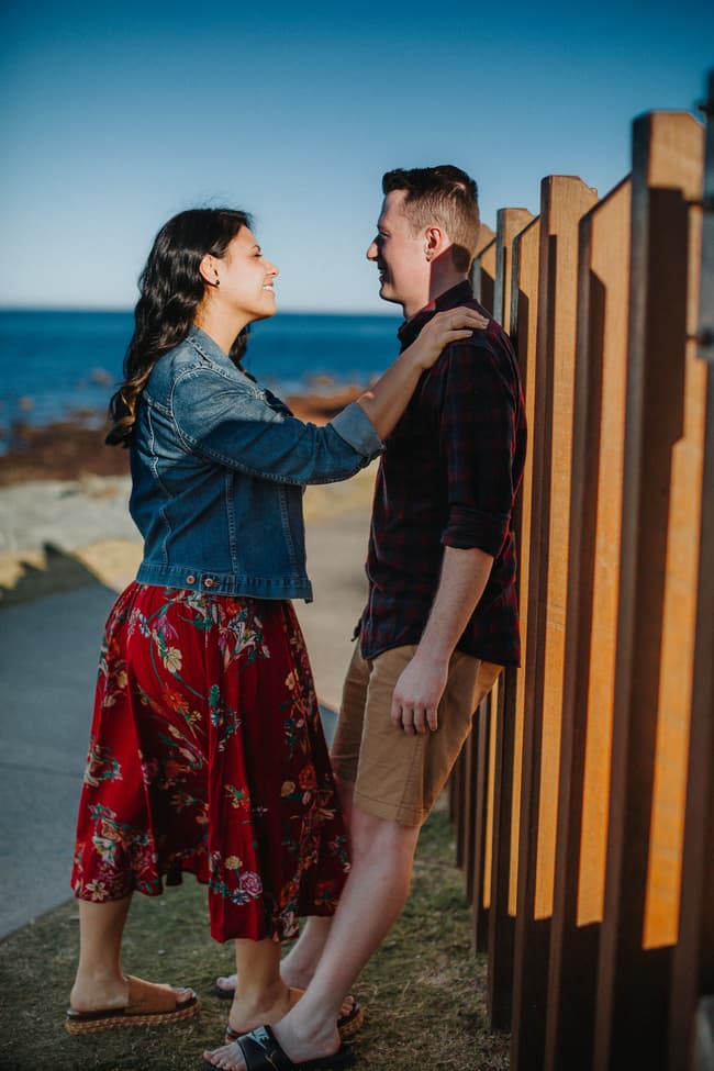 The couple stands by a wooden fence at Redcliffe, smiling at each other in warm sunlight.