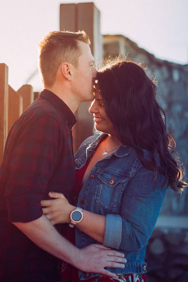 The couple embraces by a wooden fence at Redcliffe, with one kissing the other's forehead.