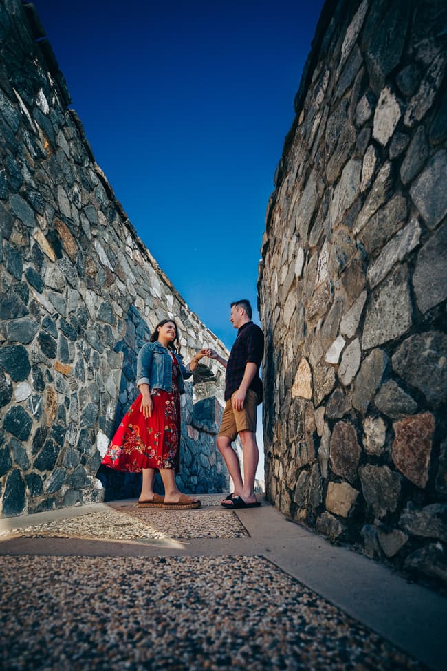 The couple stands between stone walls at Redcliffe, gazing at each other under a clear blue sky.